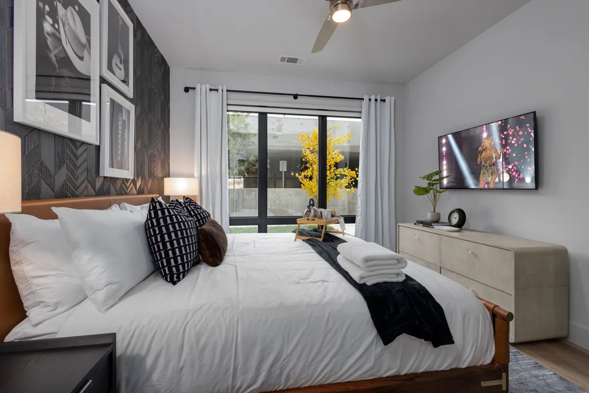 A stylish bedroom featuring a bed with crisp white linens, decorative pillows, wall art, a flat-screen television, and a window view with a tree displaying vibrant yellow foliage. The room feels airy and restful, with natural light streaming in, creating a cozy retreat.