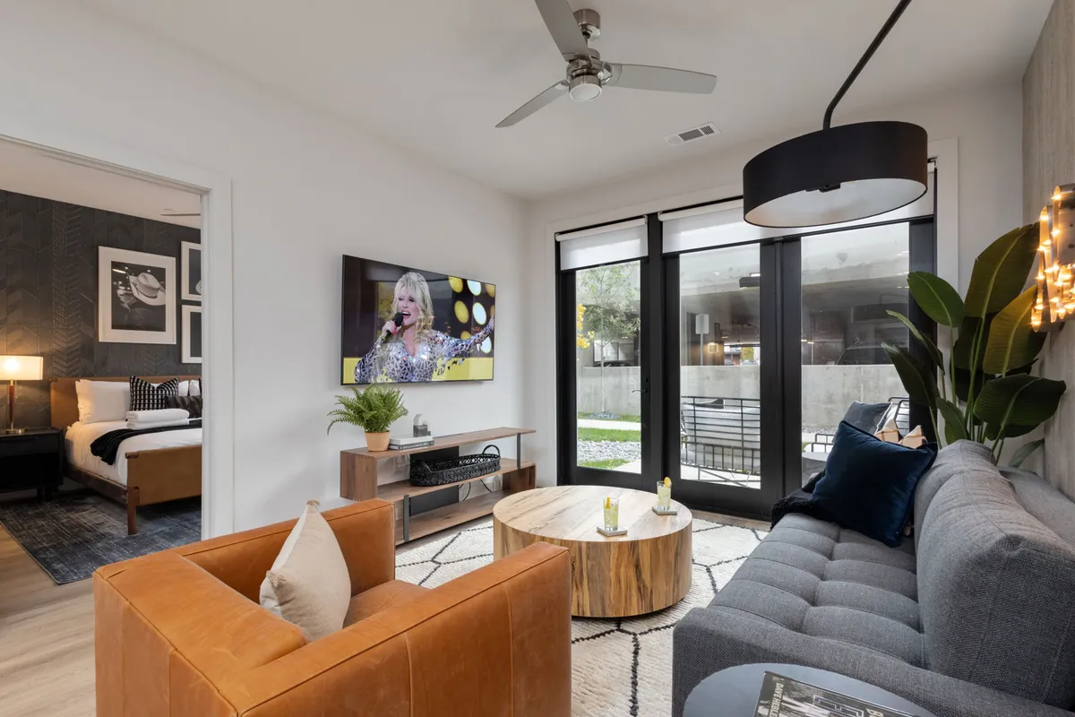 A welcoming living room with a stylish gray sofa (potentially a sleeper sofa), rustic wooden coffee table, television displaying a performance, large windows with abundant natural light, and a leather accent chair. The room sets the stage for relaxation or hosting extra guests.