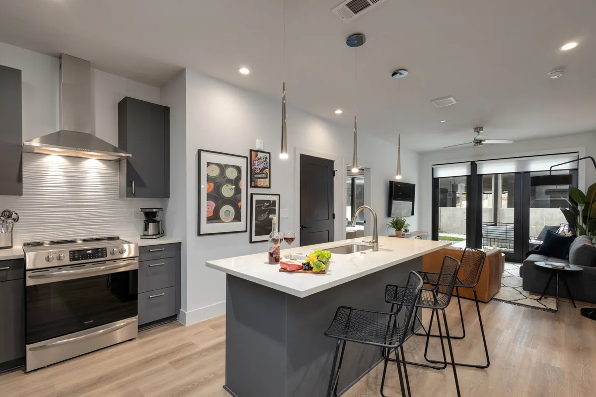 This image showcases the heart of the open-concept living area, with a modern kitchen island featuring bar stools, sleek stainless steel appliances, and contemporary wall art. The living area is visible in the background, emphasizing the seamless flow between kitchen and social spaces.