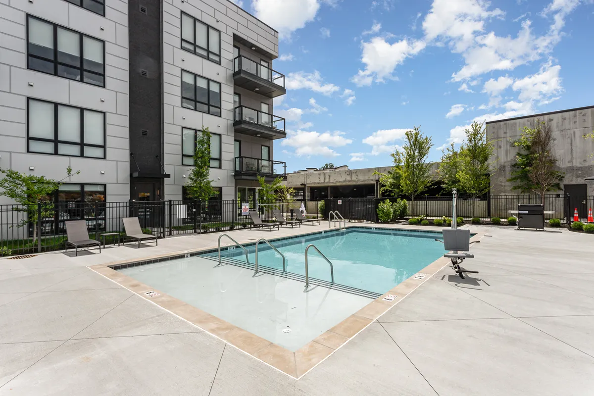 An exterior pool scene highlighting the swimming pool surrounded by lush greenery, with balconies of the building overlooking the water. Guests are shown sunbathing, swimming, and socializing, evoking a cheerful, vibrant community feel.