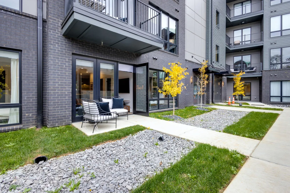 A tranquil outdoor seating area featuring modern patio furniture, surrounded by vibrant yellow autumn trees and a gravel pathway. Large glass doors hint at a seamless connection between indoor and outdoor living, while the setting sun casts a warm, inviting glow over the space.