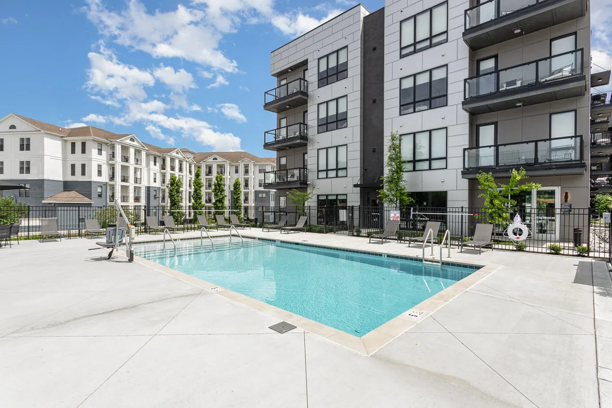 An exterior perspective of the apartment community’s swimming pool, featuring inviting lounge chairs, the apartment balconies above, and beautifully lit spaces ideal for relaxation and social gatherings. The scene captures both the tranquil ambiance of a quiet morning and the potential for lively pool parties at sunset.